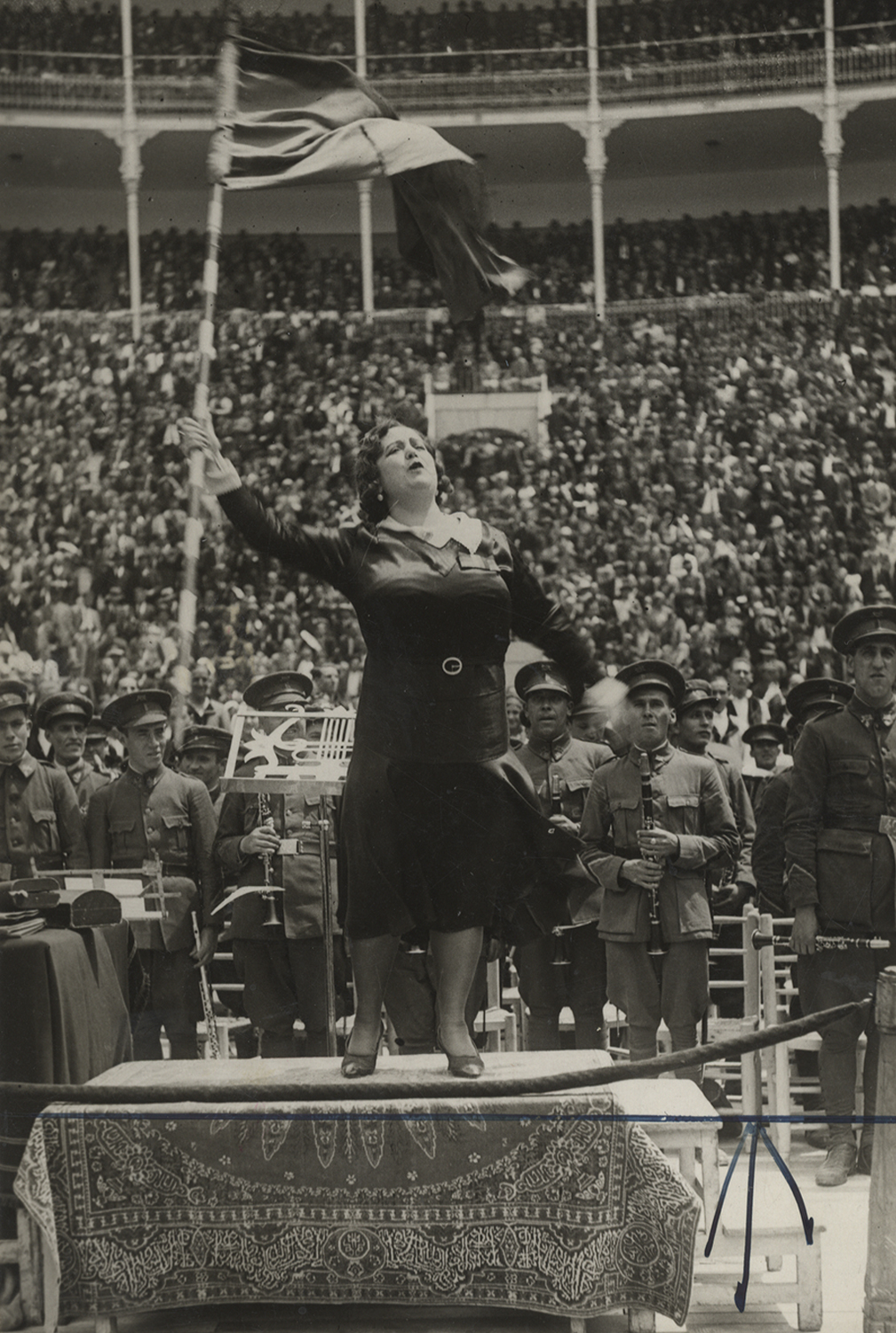 Mujer encima de una tarima, delante de músicos, ondeando una bandera