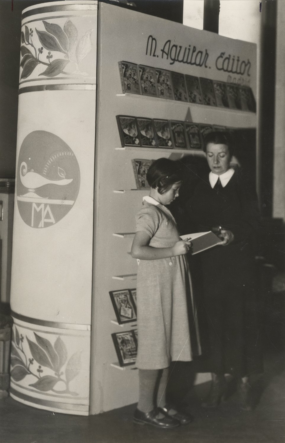 Mujer y niña con un libro delante de una estantería decorada como un libro.