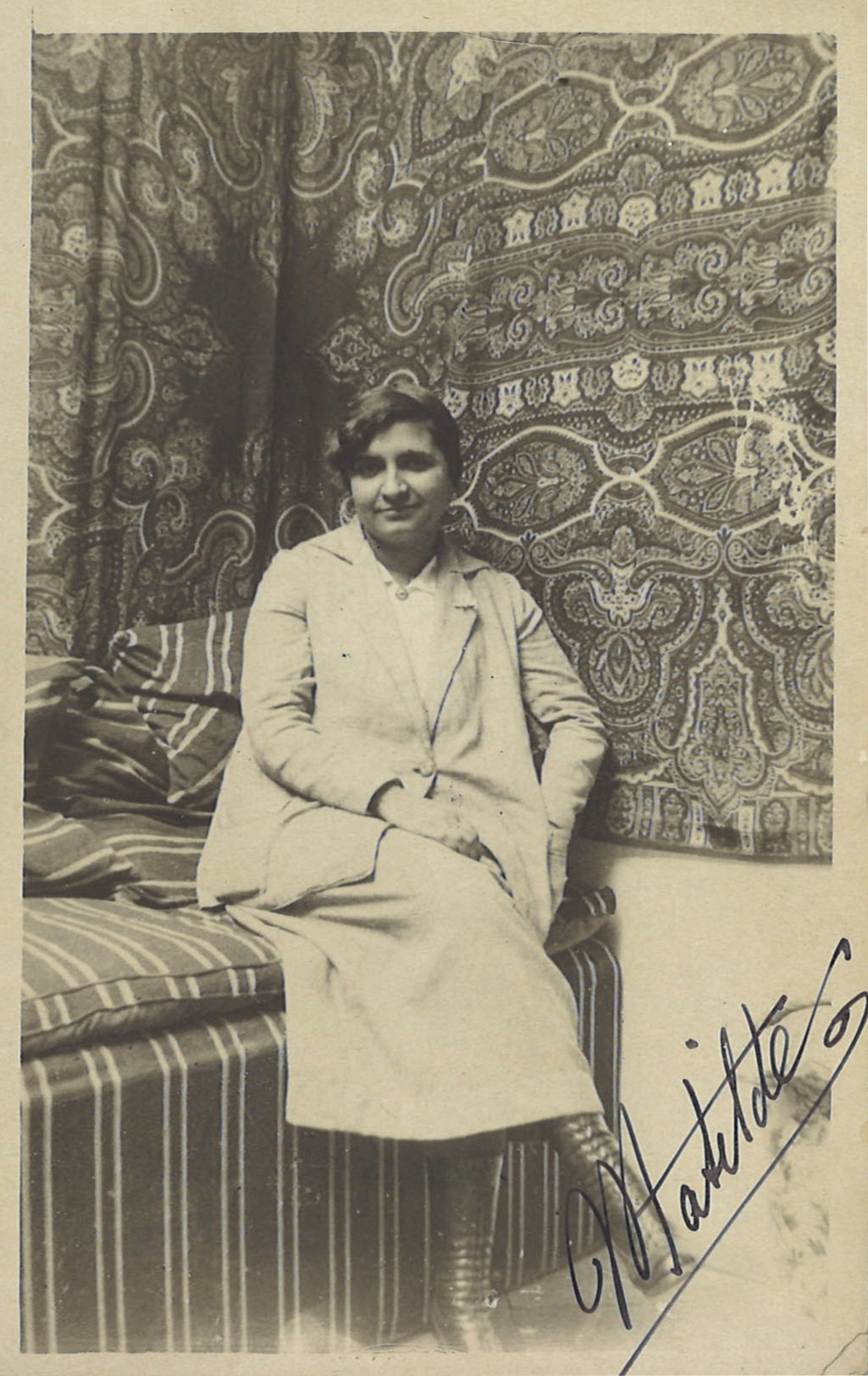 Mujer sentada en un sillon a rayas, con las piernas cruzadas, y telas en la pared detrás de ella.