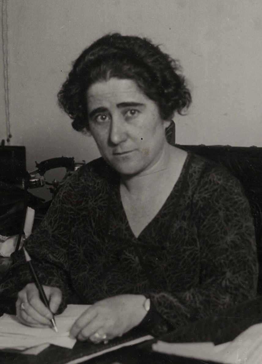 Retrato de mujer sentada escribiendo, mirando a cámara.