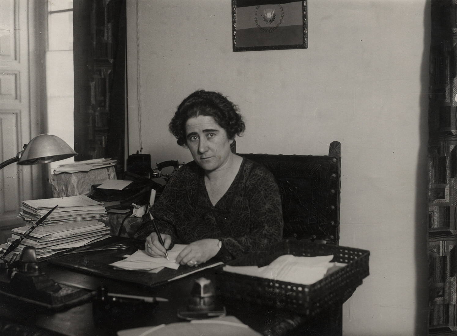 Mujer sentada en escritorio, escribiendo y mirando a cámara.