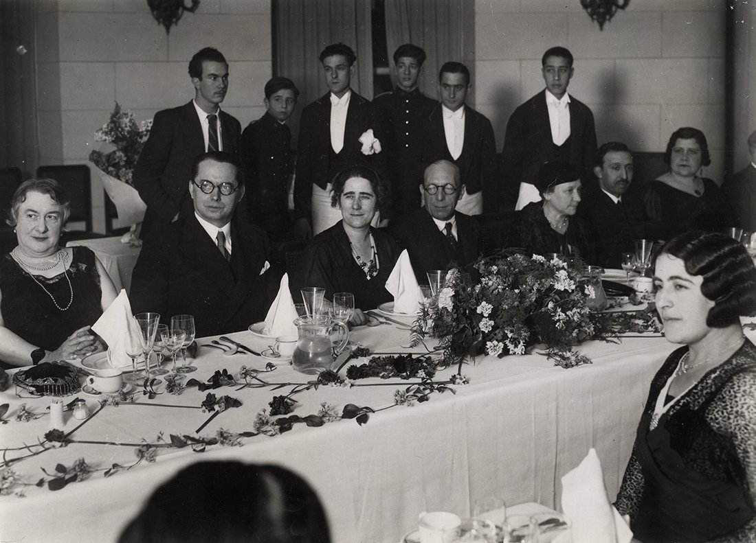 Grupo de mujeres y hombres en una mesa de comedor decorada con flores, y camareros de fondo.
