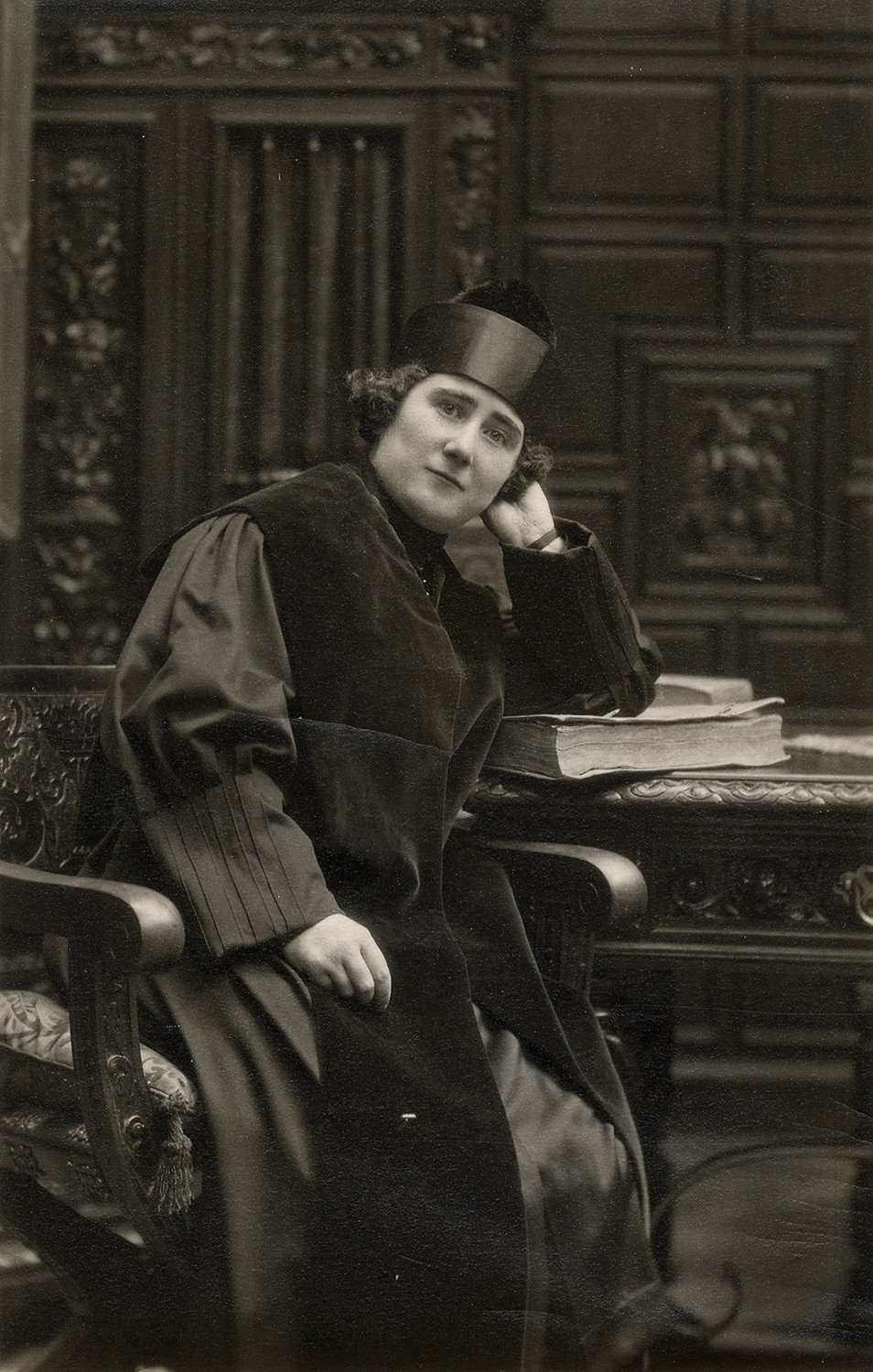 Mujer con toga de doctorado, sentada en una silla, el brazo apoyado en un libro encima de una mesa y la cabeza apoyada en la mano.