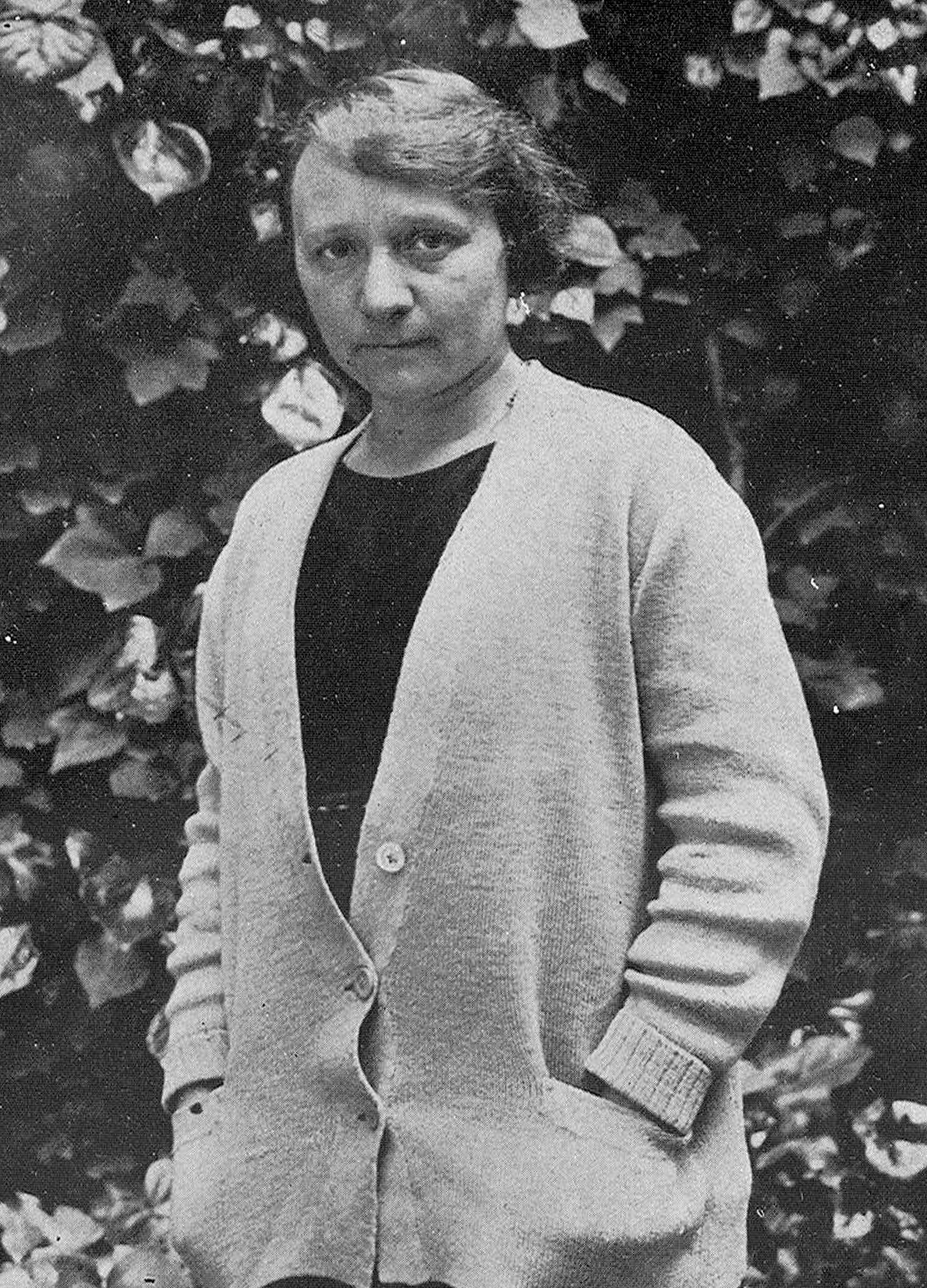 Retrato de mujer delante de hojas, con las manos en los bolsillos.