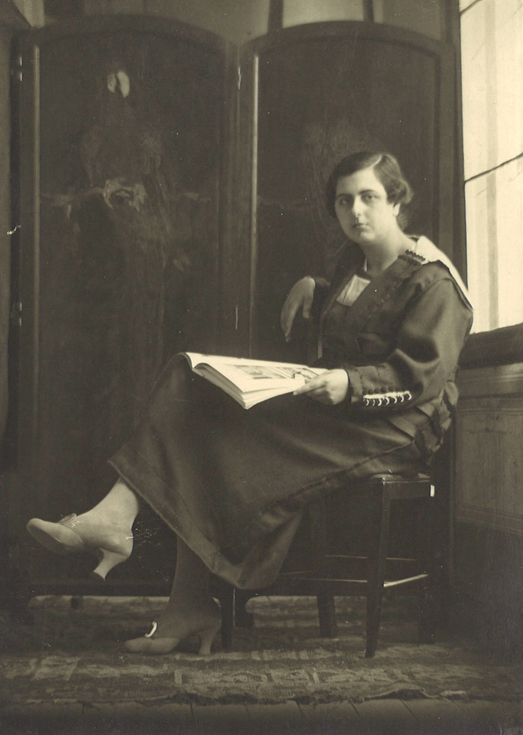 Mujer sentada con un libro en la mano, con un biombo de fondo.