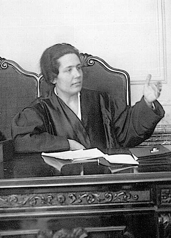 Retrato de mujer con traje de abogada, sentada en una mesa hablando.