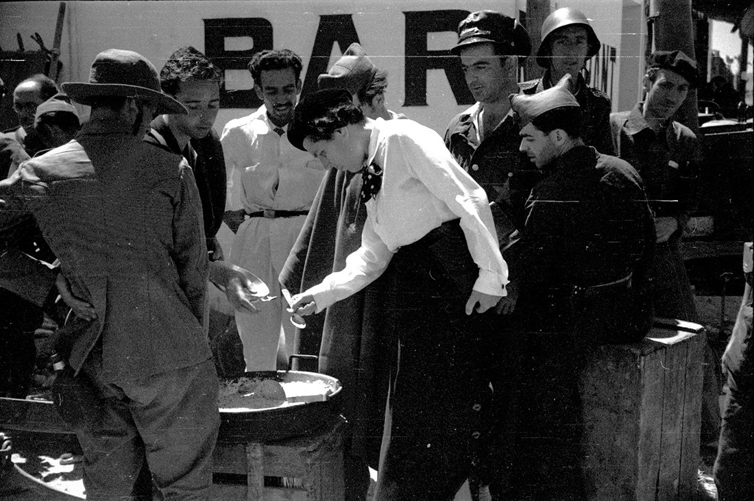 Una mujer probando comida con un grupo de hombres a su alrededor.