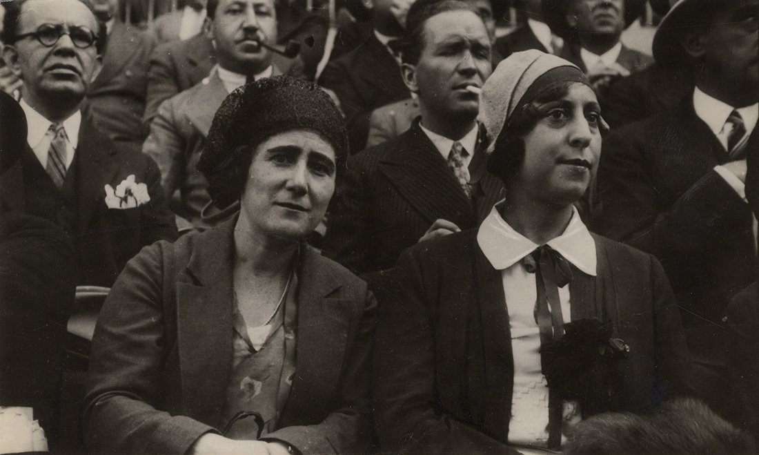 Dos mujeres en una grada, con hombres de fondo.