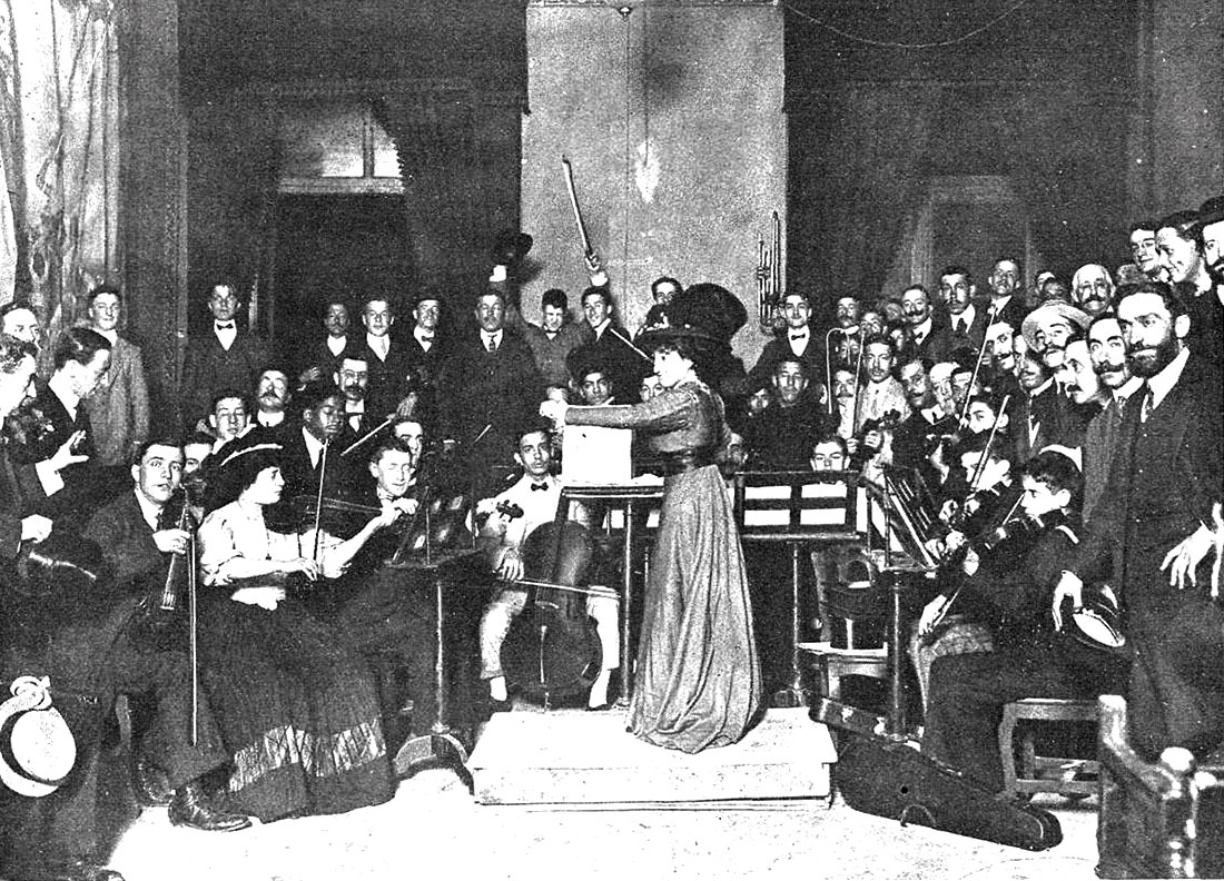 Mujer dirigiendo una orquesta y un grupo de personas mirando a cámara.