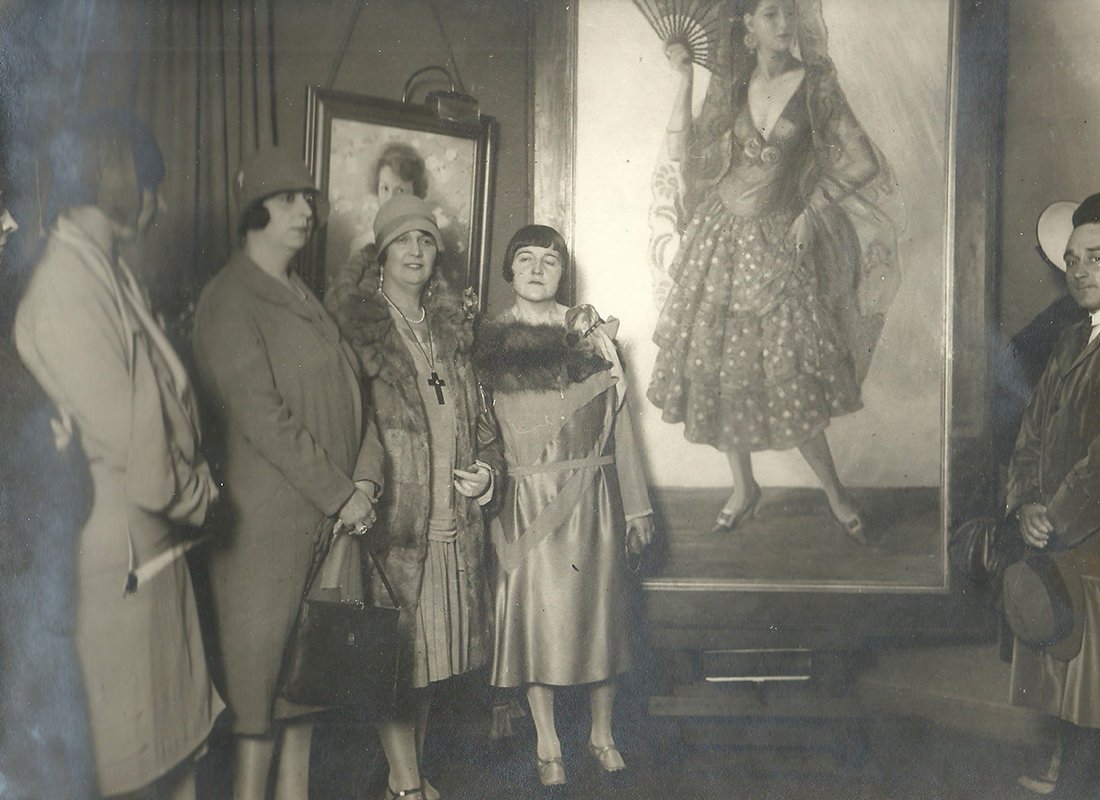 Grupo de mujeres delante de un cuadro representando una mujer con traje de sevillana y abanico.